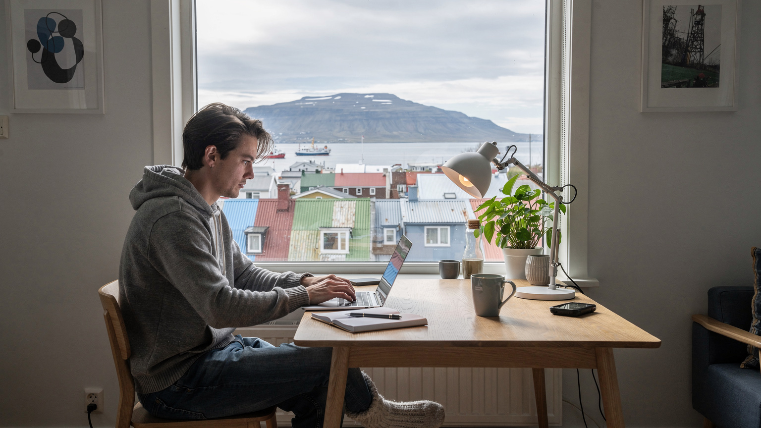En digital nomade jobber på en bærbar PC i en koselig leilighet i Reykjavik, med et stort vindu som tilbyr utsikt over byens hustak og havnen i det fjerne.