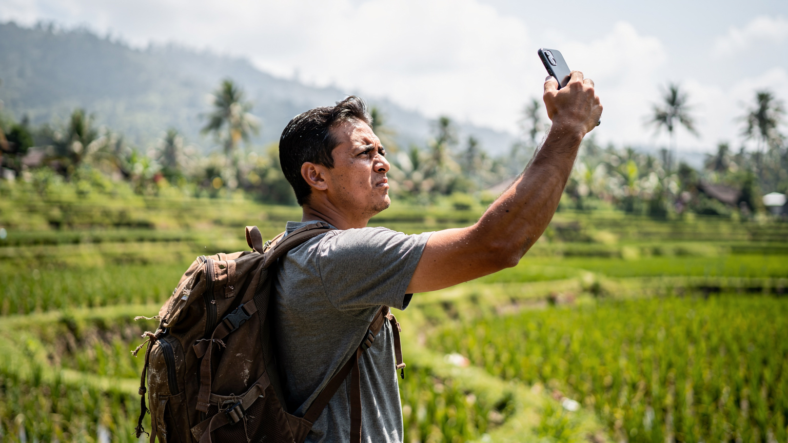 Seorang pria digital nomad sedang memegang ponselnya dengan ekspresi frustasi, mencoba mencari sinyal internet di daerah pedesaan Bali, menyoroti tantangan konektivitas di lokasi terpencil.