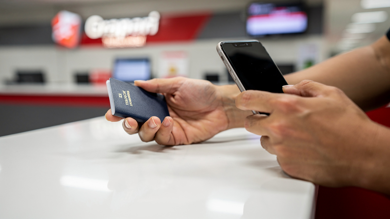 Tangan seorang traveler sedang memegang paspor dan ponsel di meja counter Grapari di bandara, menunjukkan proses registrasi atau pembelian kartu SIM.