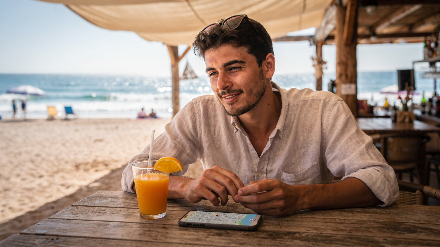 Ein junger Reisender sitzt an einem rustikalen Holztisch in einer Strandbar auf Mallorca, sein Smartphone mit einer geöffneten Karten-App liegt neben einem Glas frisch gepresstem Orangensaft. Im Hintergrund das glitzernde Meer.