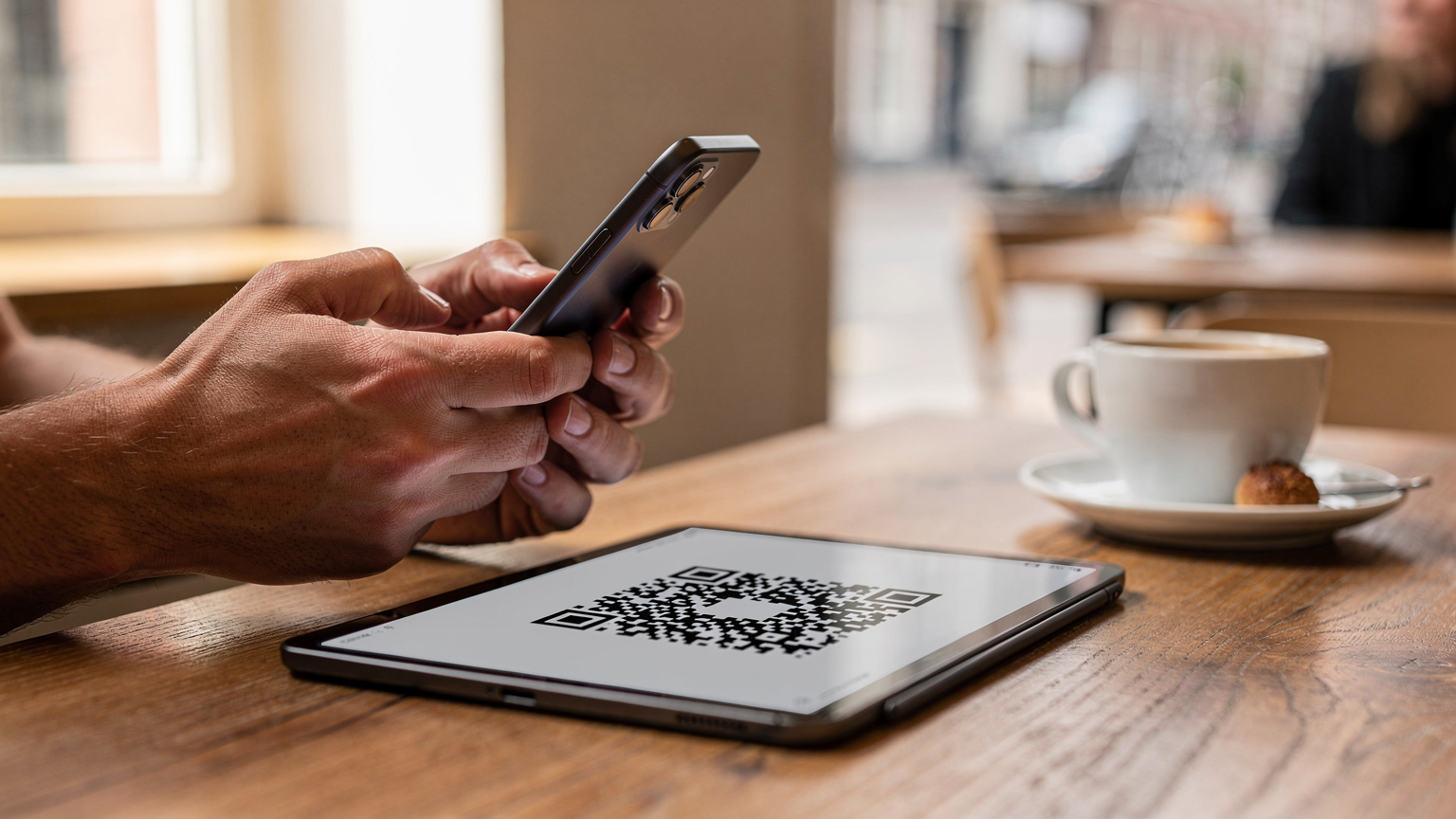 Close-up of human hands holding an iPhone 17, with a separate screen displaying a QR code for eSIM activation in a cafe setting.