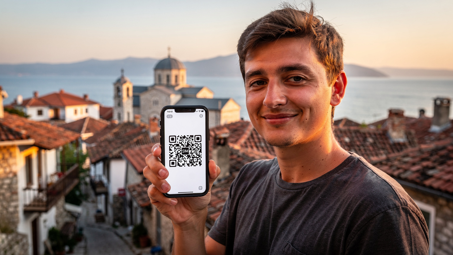 Een reiziger toont een smartphone met daarop een eSIM-activatie QR-code, met de oude stad van Ohrid en het helderblauwe meer op de achtergrond. De zonsondergang zorgt voor een warme gloed over het landschap.