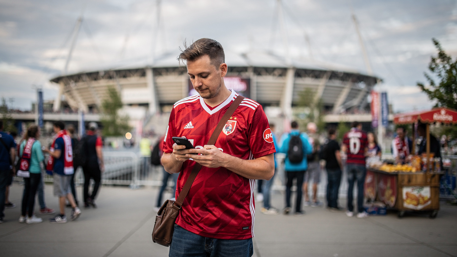 En fotballfan i en landslagsdrakt, med en mobiltelefon i hånden, navigerer et travelt stadionområde i Vancouver, Canada, med BC Place i bakgrunnen.