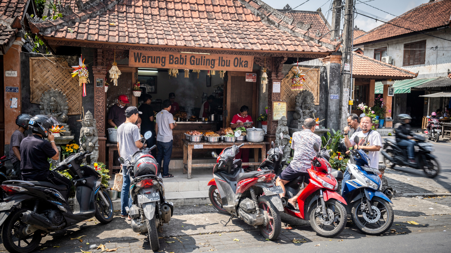 Переполненная парковка скутеров возле входа в Warung Babi Guling Ibu Oka в Убуде, Бали, с местными жителями и туристами.