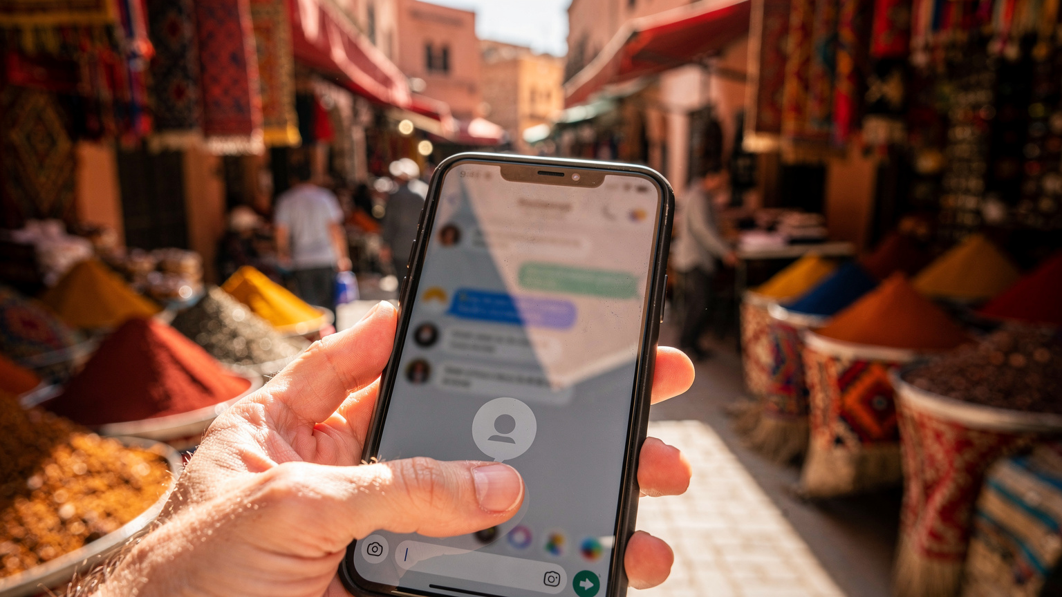 A close-up, slightly blurry shot of a smartphone screen displaying a T-Mobile support chat window, with a traveler's thumb hovering over the 'send' button. The background shows a bustling street market in Marrakech, Morocco, with vibrant textiles and spices.