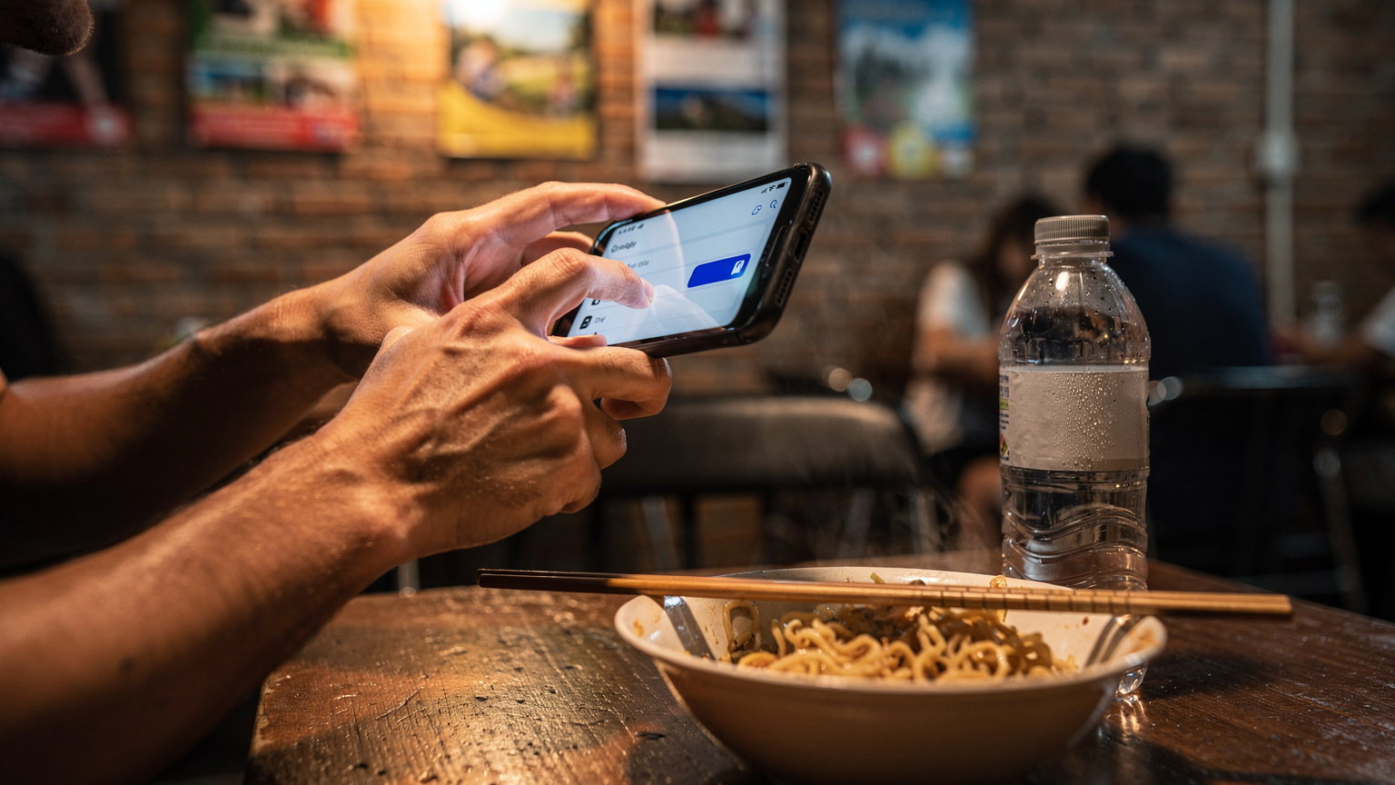 A close-up shot of a traveler's hands, specifically the thumbs, navigating the settings menu of an iPhone in a dim, cozy hostel common room. The screen shows cellular data options, with a focus on toggling eSIM profiles, while a half-eaten bowl of ramen sits nearby.