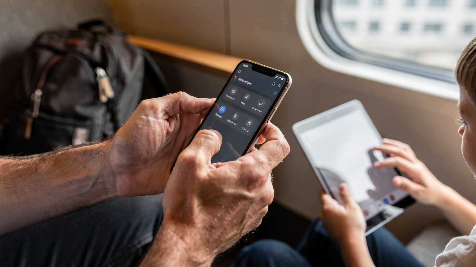 Nahaufnahme der Hände eines Vaters, der auf einem Smartphone die Einstellungen für einen mobilen Hotspot aktiviert, während im unscharfen Hintergrund ein Kind auf einem Tablet spielt.