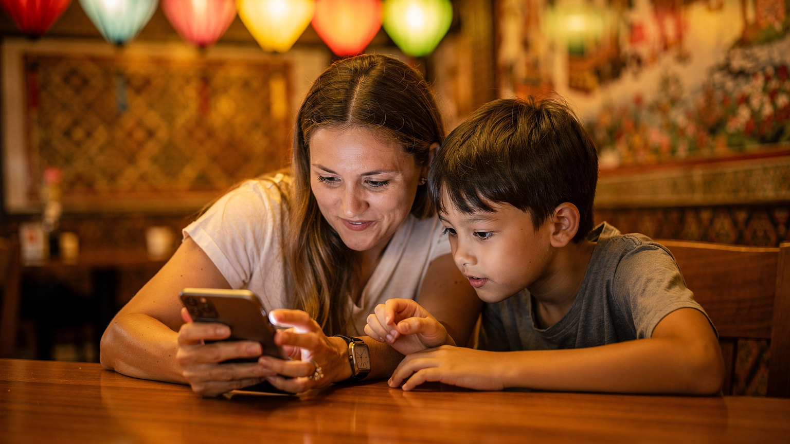Une mère montre à son enfant comment utiliser une application de contrôle parental sur son smartphone, dans un café animé de Chiang Mai, avec des lanternes colorées et des motifs thaïlandais en arrière-plan.