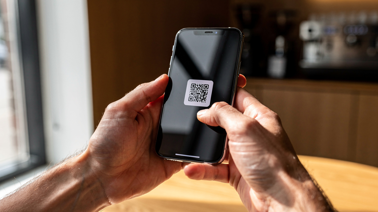Een close-up van handen die een smartphone vasthouden, waarbij een QR-code voor eSIM-installatie wordt gescand in een modern, licht café in Amsterdam-Noord.