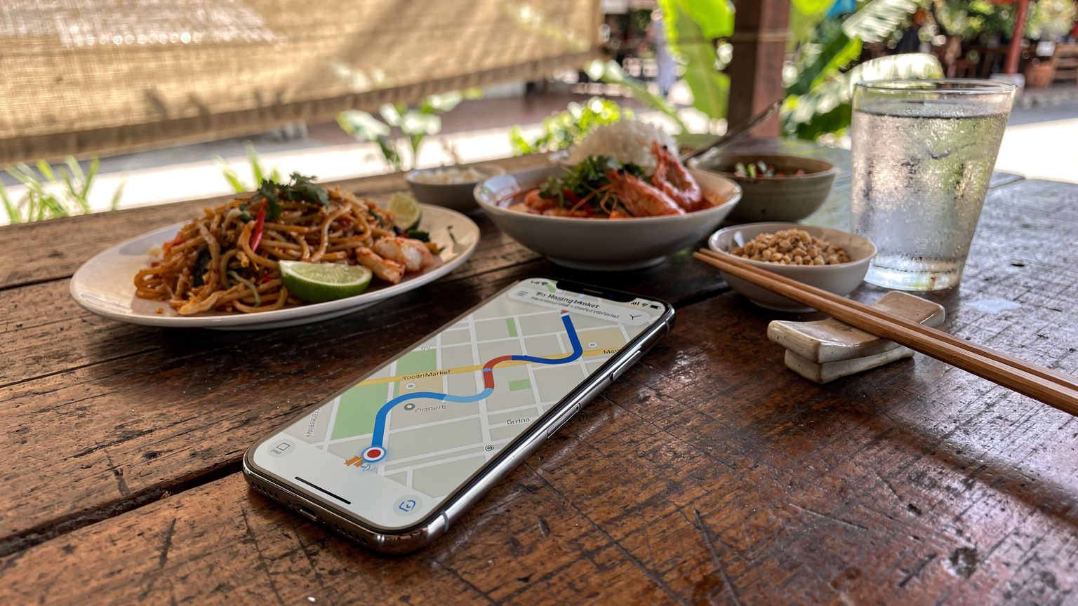 Un smartphone affichant une application de cartographie avec un itinéraire vers un marché local, posé sur une table en bois d'un restaurant à Chiang Mai, avec des plats thaïlandais colorés et des baguettes en arrière-plan.