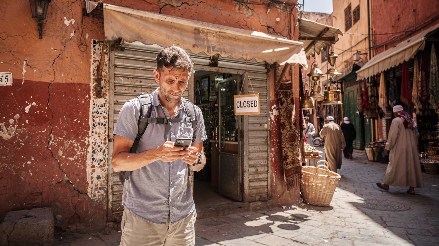 Ein Reisender steht frustriert vor einem geschlossenen Mobilfunkgeschäft in einer belebten marokkanischen Gasse, sein Smartphone in der Hand, was die Schwierigkeiten bei der Beschaffung einer lokalen SIM-Karte symbolisiert.