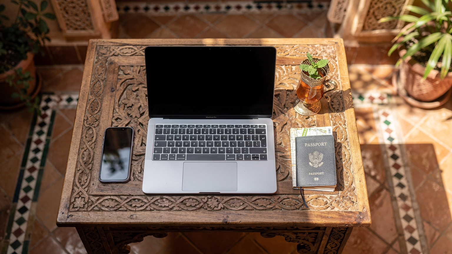 Ein Reisender entspannt in einem traditionellen marokkanischen Riad, sein Smartphone liegt neben einem Laptop und einer Tasse Minztee auf einem kunstvoll geschnitzten Holztisch, was die Balance zwischen Arbeit und Entspannung unterstreicht.