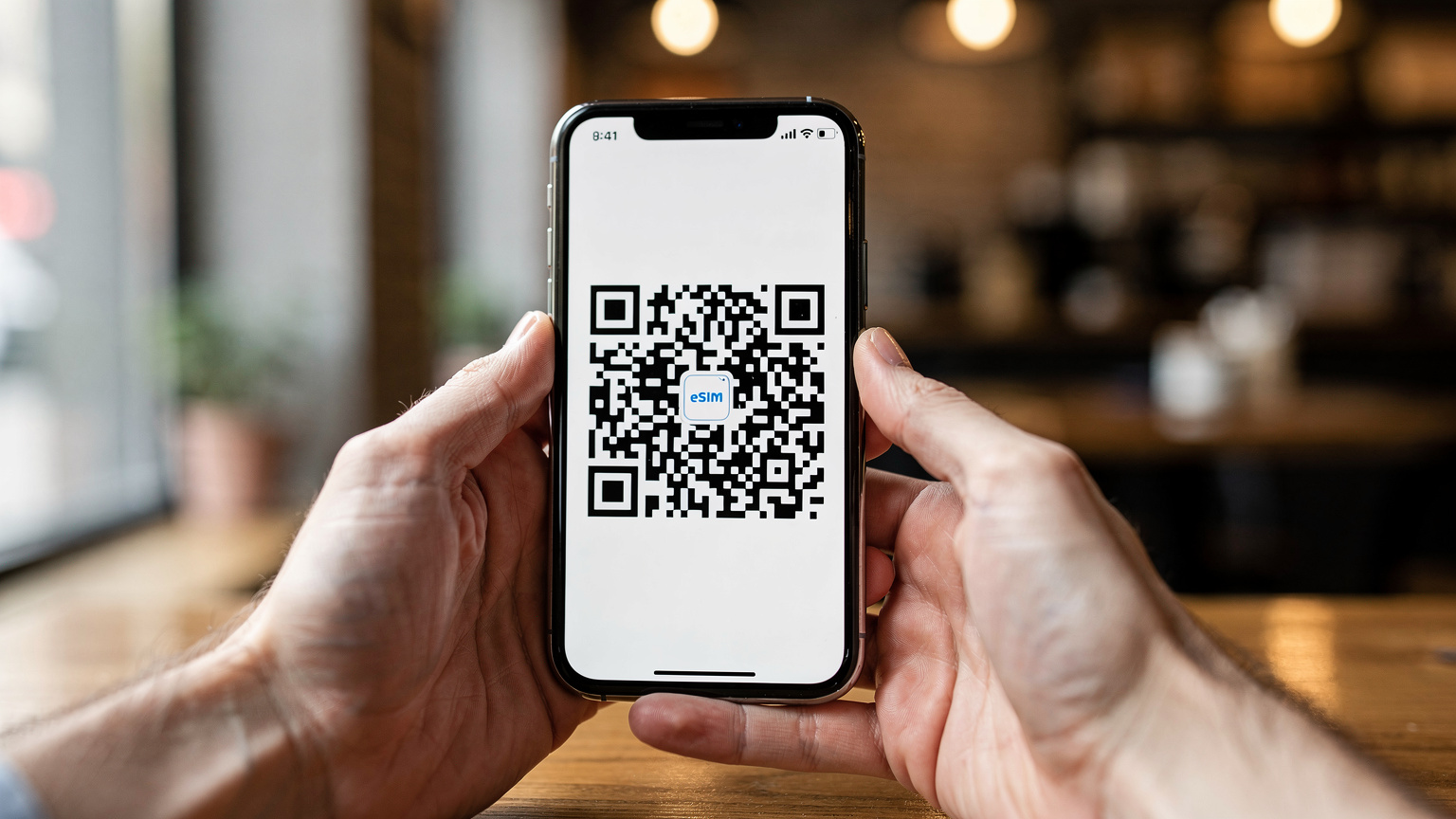 Крупний план рук, що тримають смартфон, на екрані якого відображається QR-код для активації eSIM, на задньому плані розмитий інтер'єр кафе.