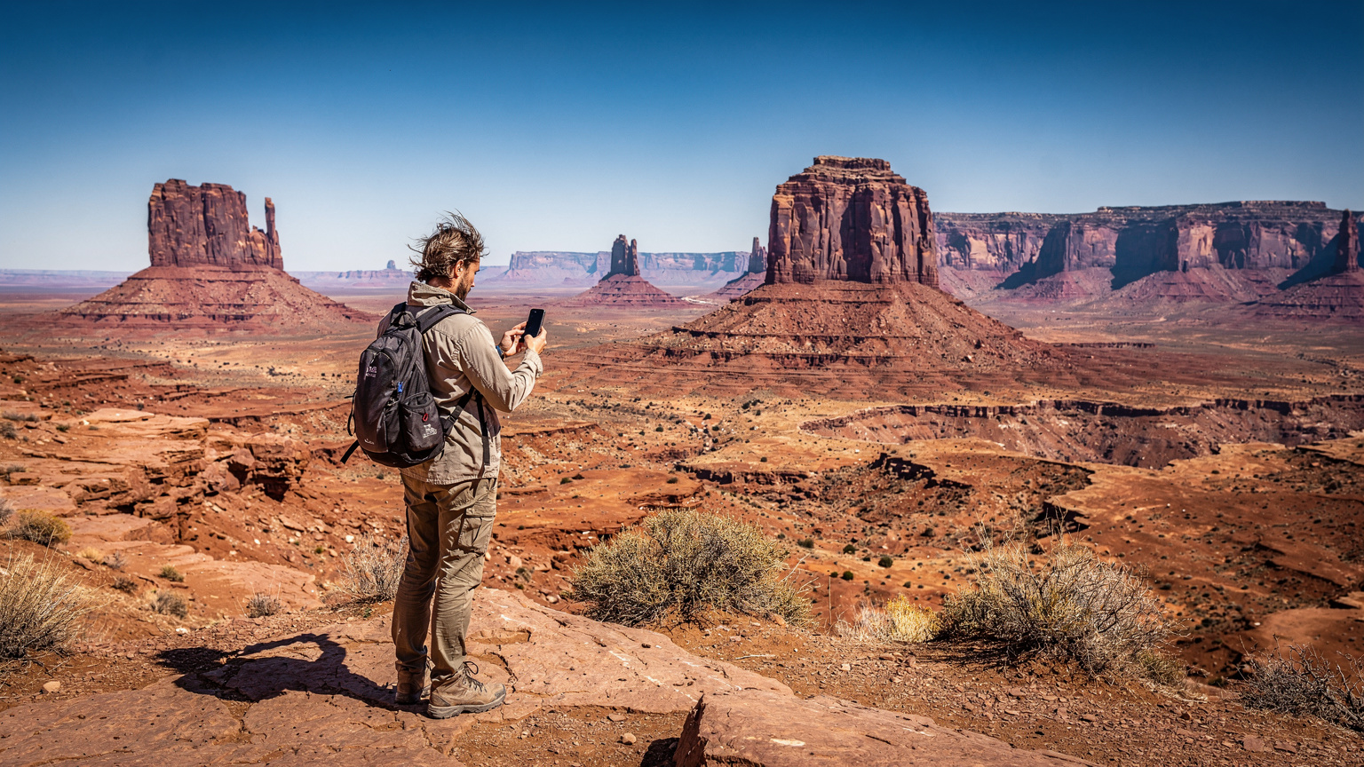 A lone traveler stands at a scenic overlook in Monument Valley, Arizona, looking at their smartphone with a concerned expression, symbolizing the need for reliable network signal in remote areas.
