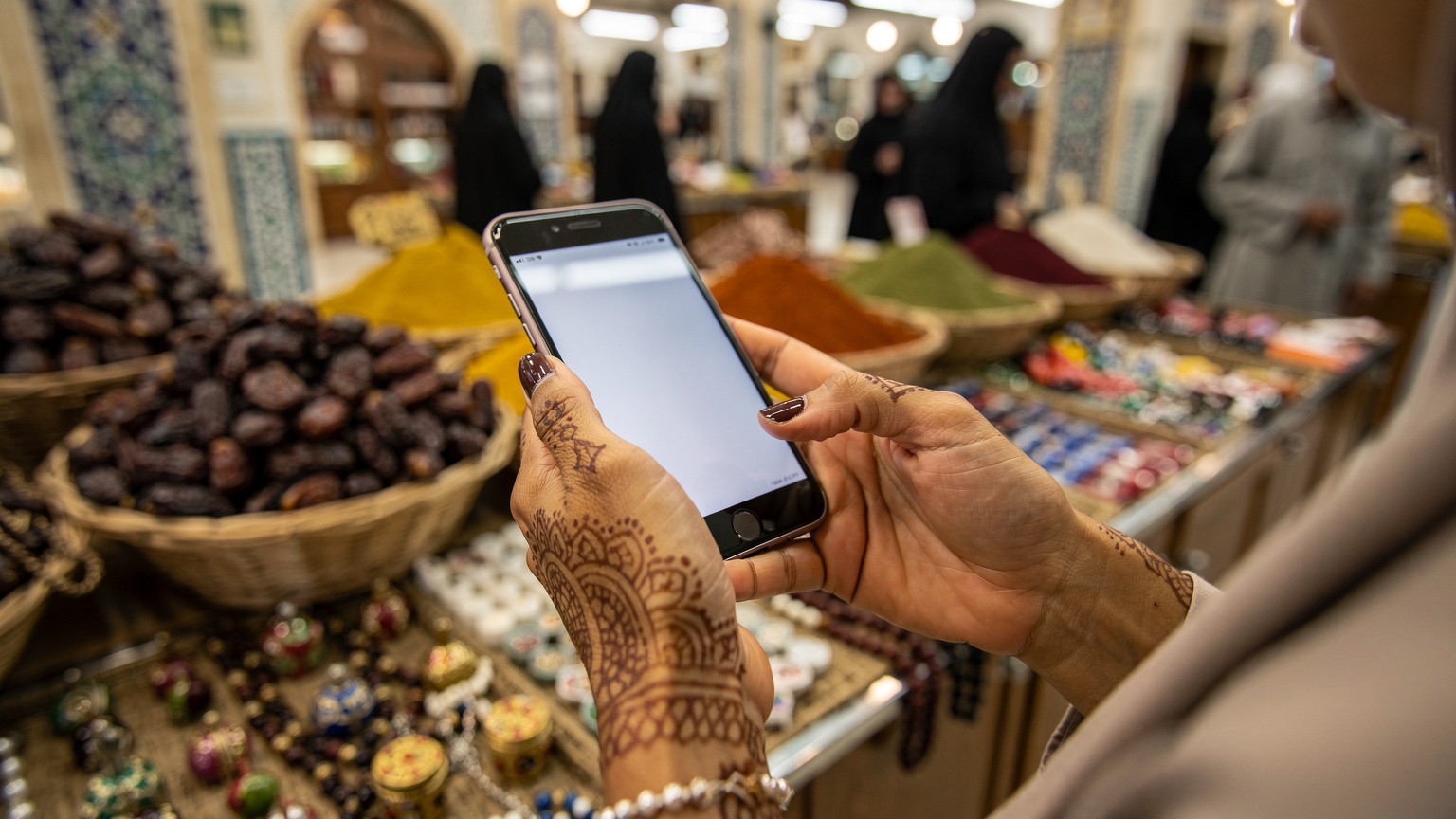 Tangan seorang jemaah haji yang sedang memegang telefon pintar dengan latar belakang pasar di Madinah, menunjukkan aktiviti membeli-belah atau mencari maklumat menggunakan sambungan eSIM.
