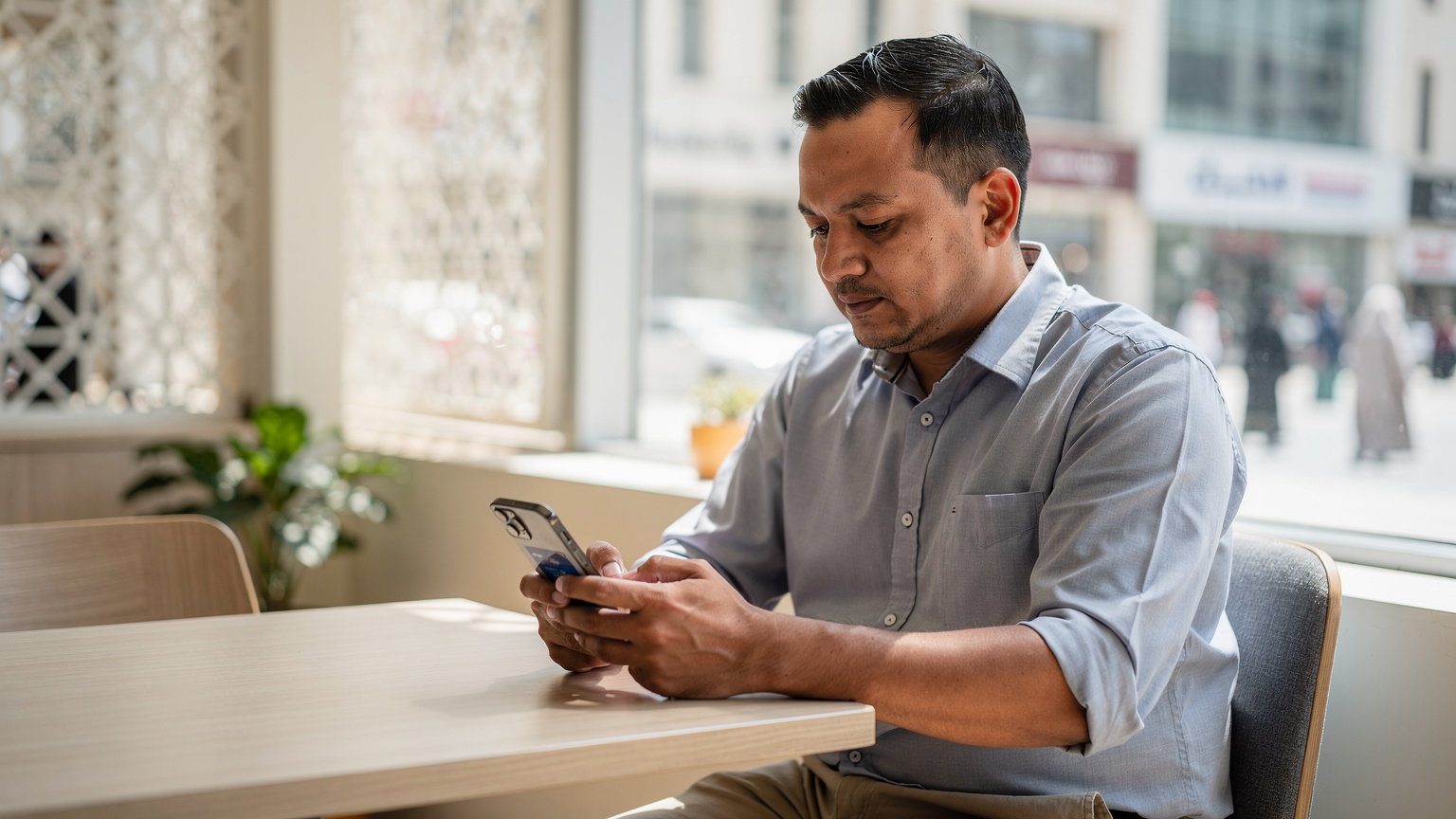 Seorang jemaah lelaki Malaysia yang lebih muda sedang duduk di kafe moden di Jeddah, memegang telefon pintar dan mengimbas kod QR untuk mengaktifkan eSIMnya, menunjukkan kemudahan persediaan sebelum keluar ke bandar.