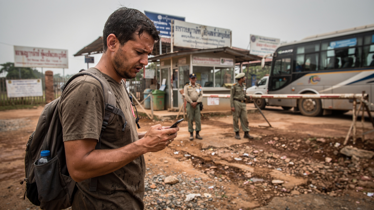 Een reiziger staat verdwaasd en gefrustreerd bij een landsgrenscontrolepunt in Zuidoost-Azië, met een ongeruste uitdrukking terwijl hij naar zijn telefoon kijkt, worstelend met connectiviteit na het oversteken van de grens.