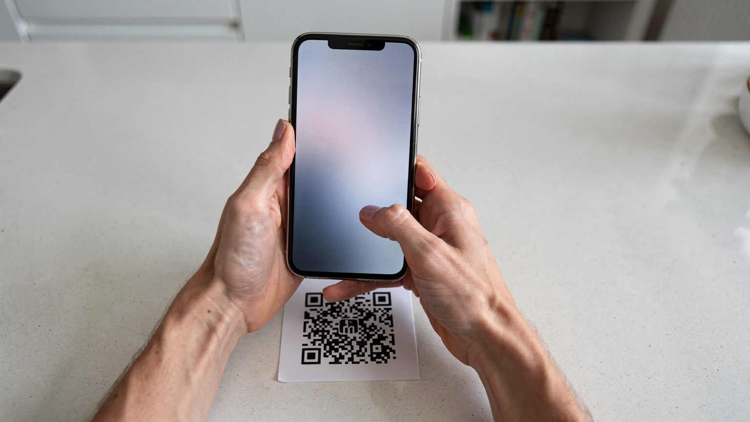 Mãos de um adulto seguram um smartphone sobre um balcão de cozinha claro, digitalizando um código QR impresso num folheto. O fundo desfocado sugere uma casa limpa e moderna.