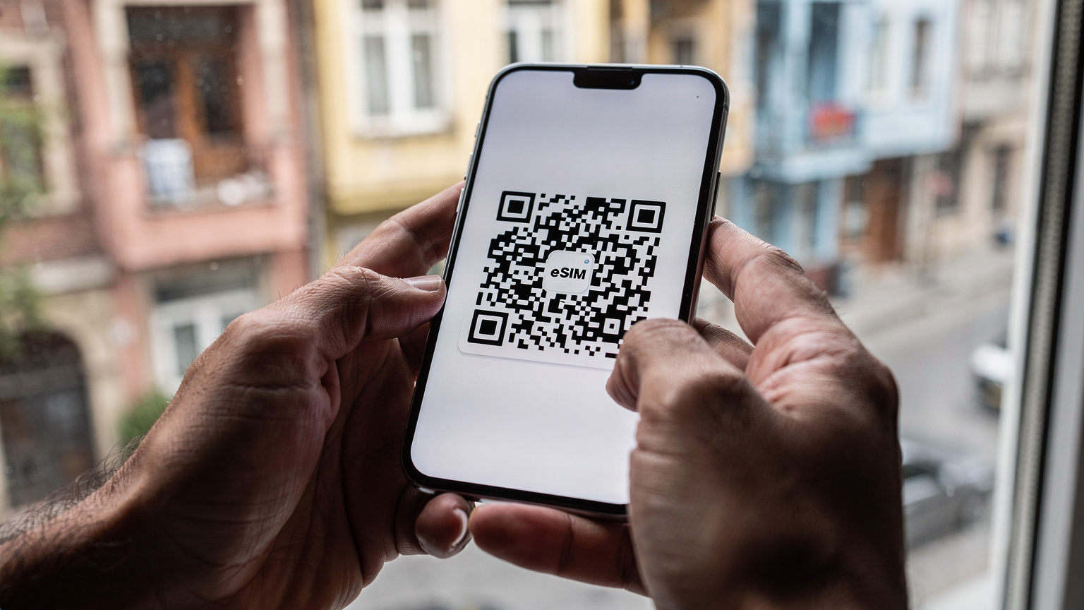 Близък план на ръце, държащи смартфон, който показва QR код за активация на Cellesim eSIM, на фона на прозорец с изглед към историческа улица в Истанбул.