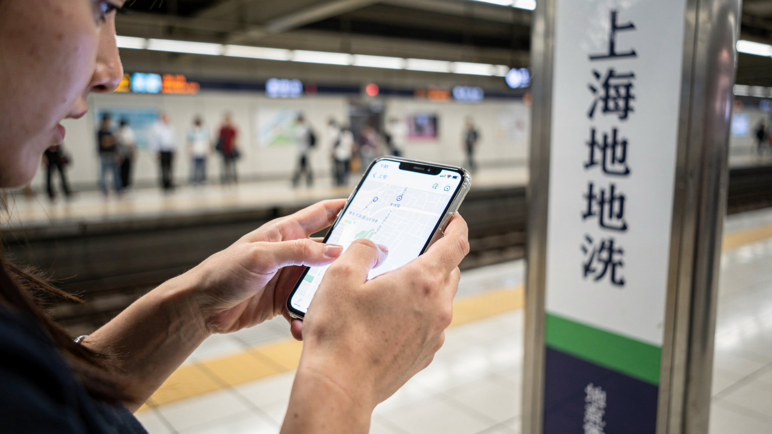 Một phụ nữ đang dùng ngón tay cái để điều hướng ứng dụng bản đồ trên iPhone của mình, đứng cạnh biển chỉ dẫn bằng tiếng Trung tại ga tàu điện ngầm Thượng Hải.