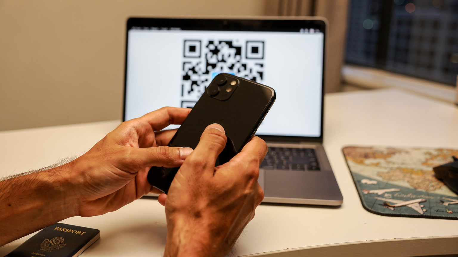 एक व्यक्ति अपने iPhone से Cellesim के eSIM QR कोड को स्कैन कर रहा है, जो यात्रा के लिए डेटा सक्रिय कर रहा है, एक लैपटॉप स्क्रीन पर QR कोड दिख रहा है।