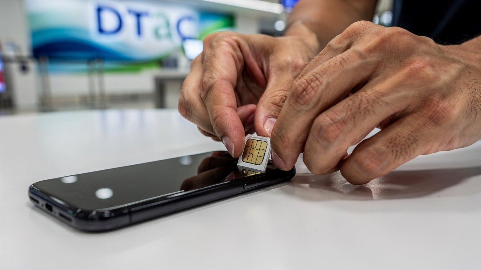 A close-up of a traveler's hands carefully inserting a small physical SIM card into the SIM tray of a smartphone, at a brightly lit mobile phone store counter in a Thai airport.