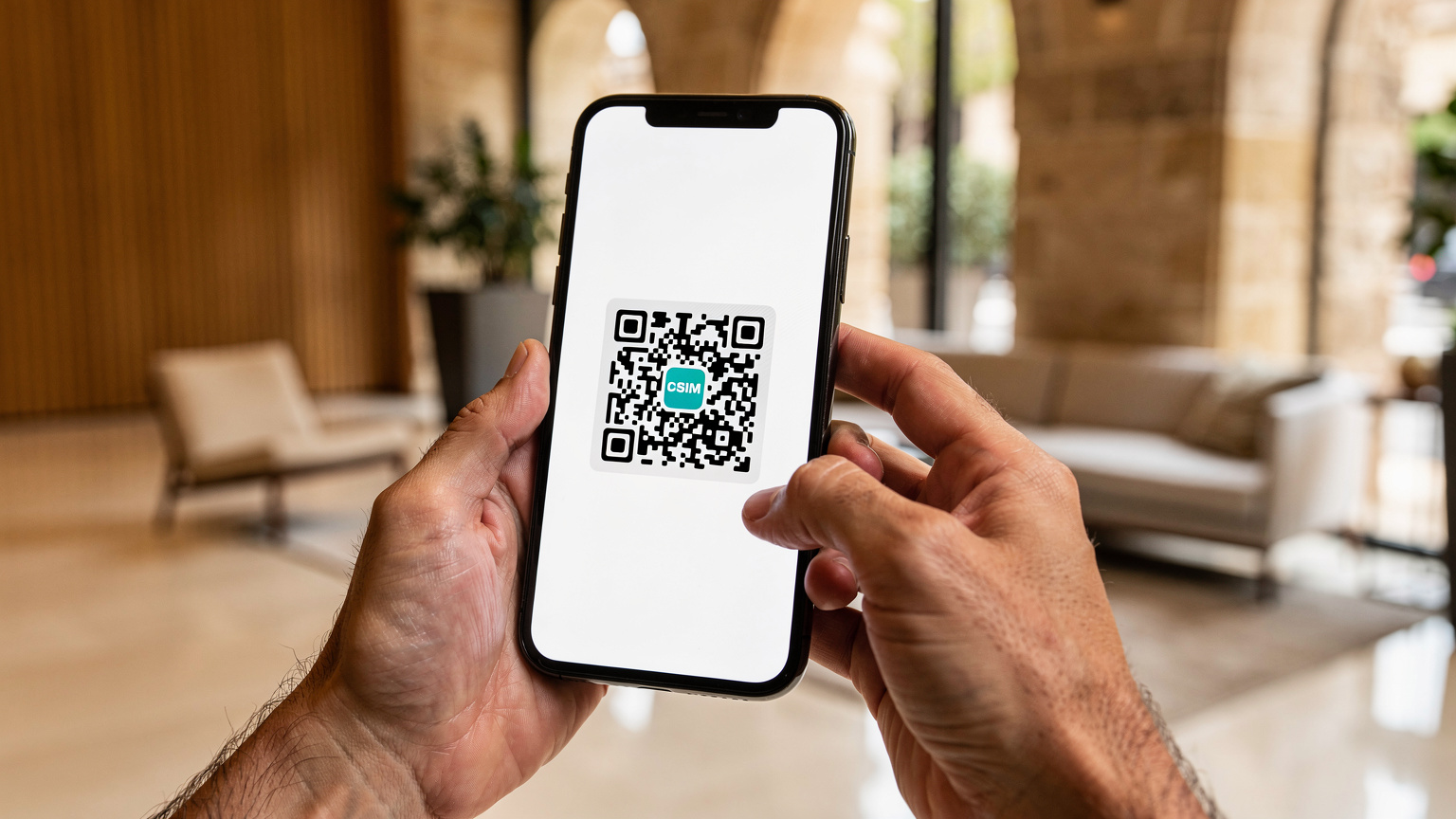 En närbild på händerna på en man i 30-årsåldern som håller upp en smartphone med en QR-kod på skärmen, redo att skannas för eSIM-aktivering, med en suddig bakgrund av en modern hotellobbyn i Barcelona.