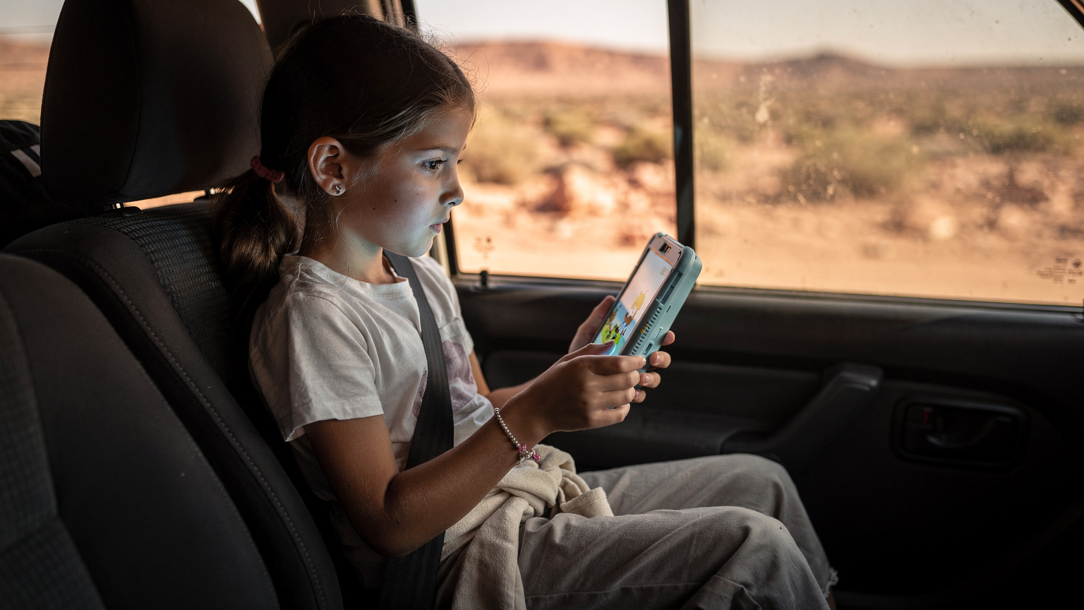 Ein 10-jähriges Mädchen auf der Rückbank eines Geländewagens in Marokko, konzentriert auf ein Tablet, das eine Zeichentrickserie abspielt, während das Fahrzeug über eine staubige Piste fährt.