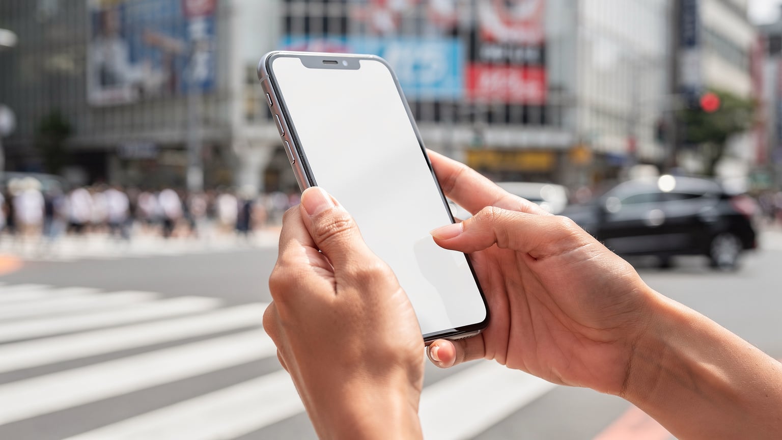 Tangan seorang wanita memegang ponsel dengan aplikasi Cellesim terbuka, menampilkan pilihan paket eSIM untuk Jepang, sementara latar belakangnya buram menampilkan pemandangan Shibuya Crossing yang ramai.