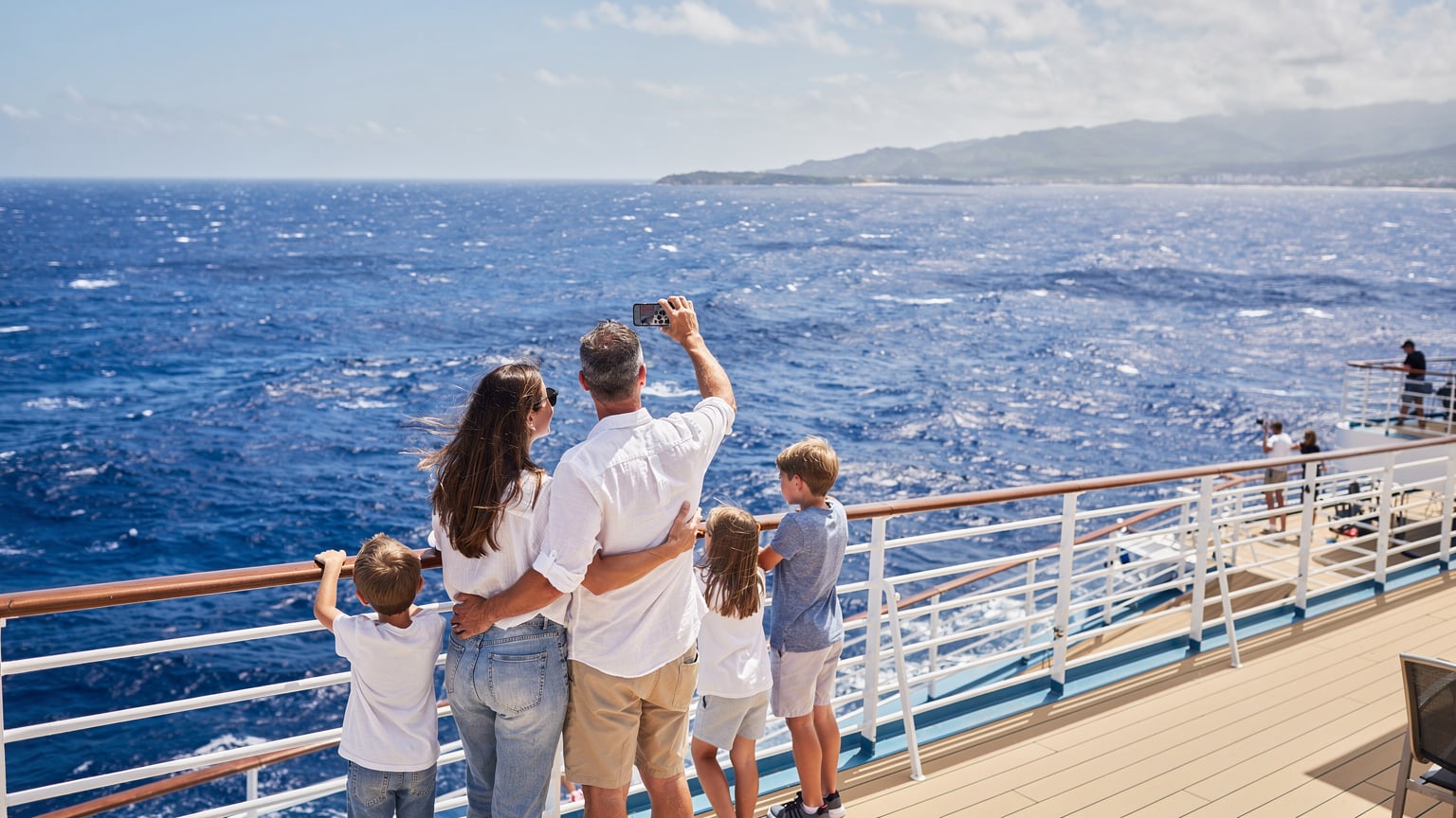A family with two children, aged 7 and 11, standing on the deck of a cruise ship, looking out at the ocean, with the father holding an iPhone 15, checking for signal as they near a port.