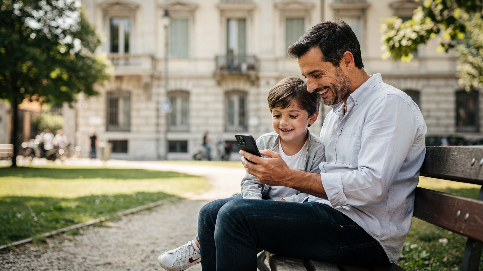 Un padre e un figlio seduti su una panchina in un parco a Milano, osservano un telefono insieme, con l'architettura classica milanese sullo sfondo e luce del primo pomeriggio.