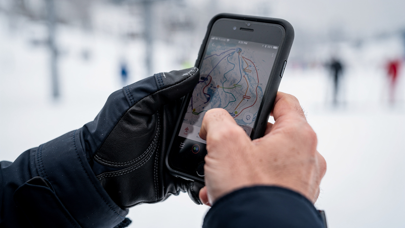 Eine Nahaufnahme zeigt Hände, die ein Smartphone halten, auf dem eine Karten-App mit Skigebieten geöffnet ist, während die Person einen Skihandschuh trägt.