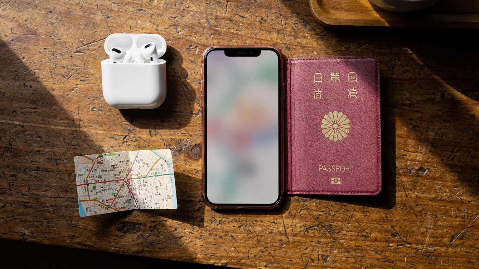 An editorial flat-lay showing a Japanese passport, a smartphone displaying a map app, a pair of wireless earbuds, and a small, folded paper map of Tokyo, all arranged on a wooden table in a cafe.