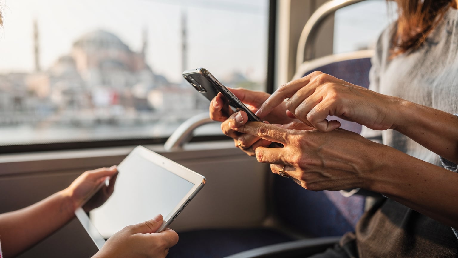 İstanbul'da bir tramvayda, kalabalık içinde bir annenin eli, çocuğunun elindeki tablete hotspot bağlantısı kurmak için kendi telefonunda ayar yaparken. Arka planda Tarihi Yarımada'nın silüeti hafifçe seçiliyor.