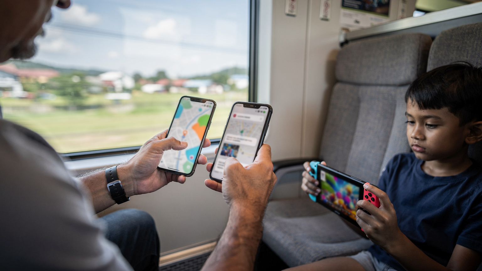 Tangan seorang ayah sedang memegang dua smartphone, satu menampilkan peta digital George Town dan yang lain notifikasi penggunaan data, di samping anaknya yang berusia 9 tahun asyik bermain game di Nintendo Switch. Latar belakangnya adalah interior kereta KTM Antarabandar yang bergerak di Malaysia.