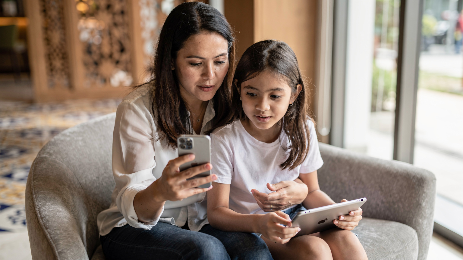 Seorang ibu menunjukkan pengaturan Screen Time di iPhone-nya kepada anak perempuannya yang berusia sekitar 10 tahun, yang sedang memegang iPad. Mereka duduk di sofa yang nyaman di lobi hotel modern di Kuala Lumpur.