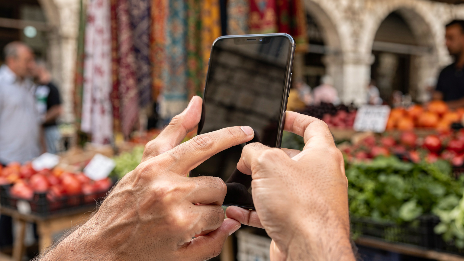 Närbild på en hand som håller en smartphone och navigerar genom inställningar för att installera ett eSIM, med en oskarp bakgrund av en traditionell albansk marknad.