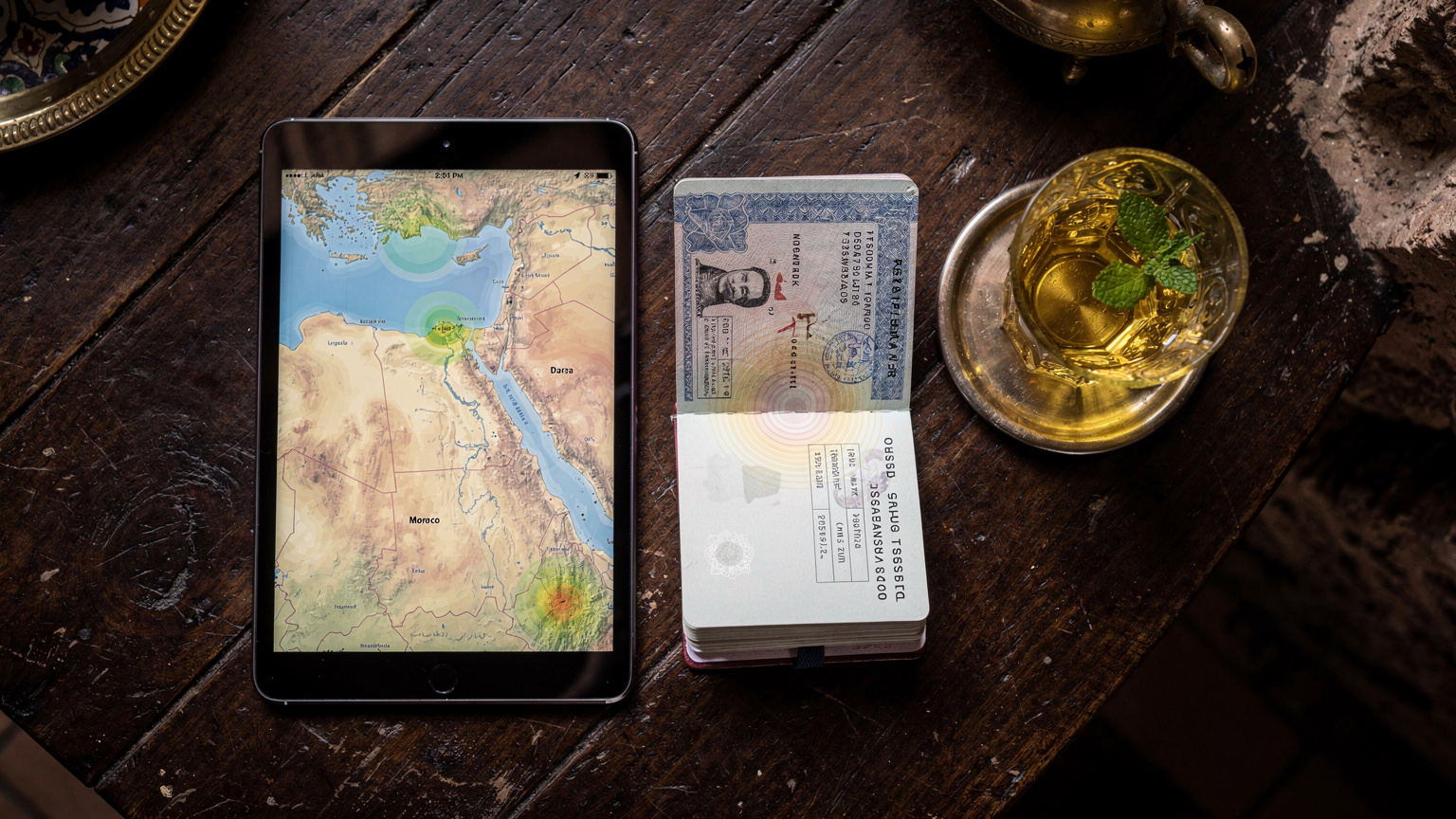 Un mapa digital de Marruecos en un tablet con superposiciones de cobertura de red, junto a un pasaporte y un vaso de té de menta en una mesa de madera de un café en Fez.
