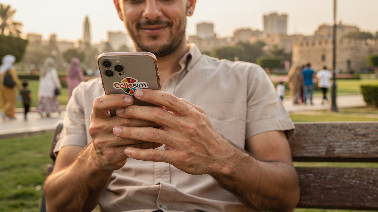 Un primo piano delle mani di un uomo italiano che tiene il suo smartphone con un adesivo colorato sul retro che dice "Cellesim", mentre digita un messaggio con un'espressione rilassata, seduto su una panchina in un parco pubblico a Il Cairo, con la gente del posto che passeggia sullo sfondo.