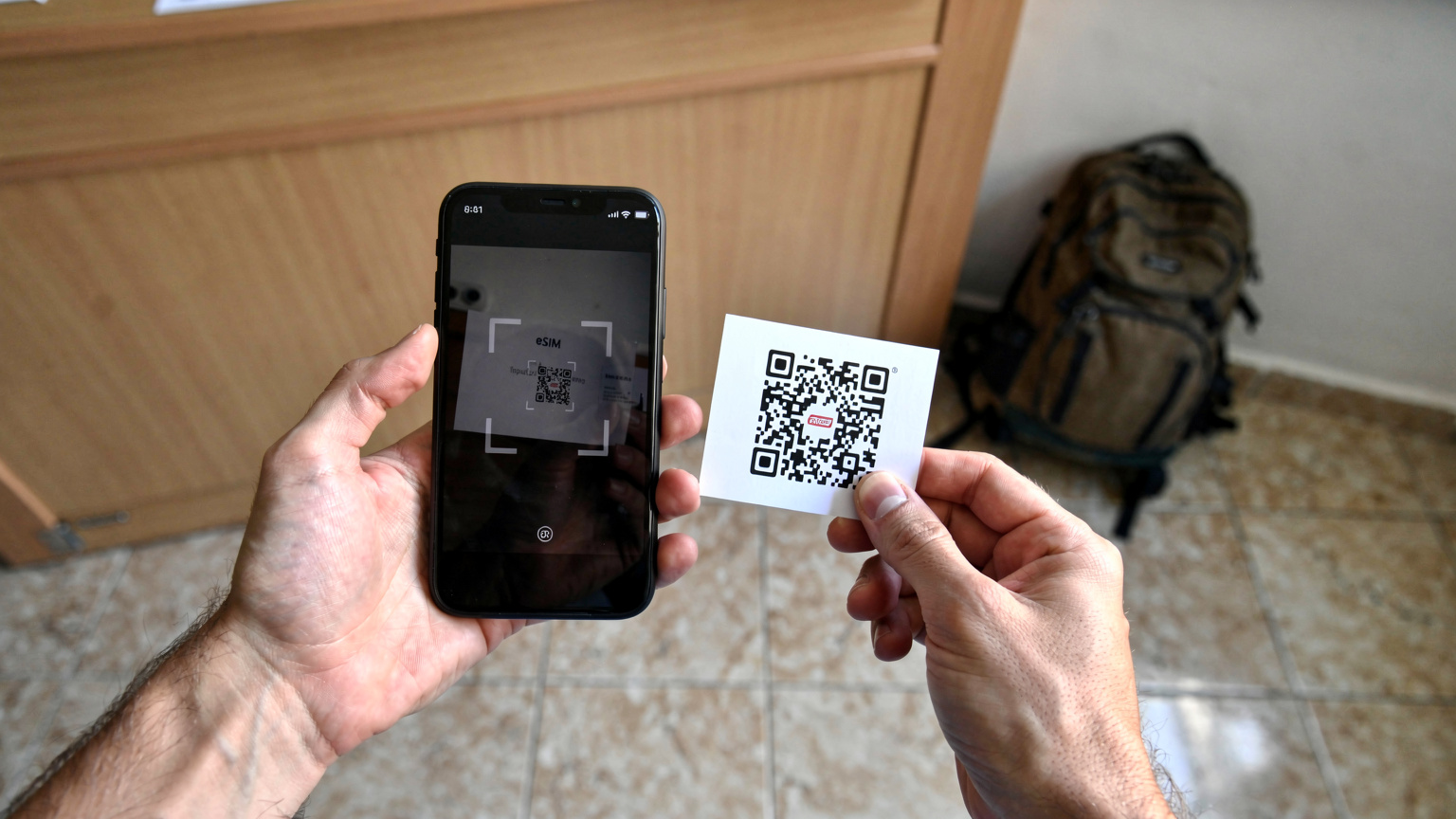 Ένα ζευγάρι χέρια κρατά ένα smartphone με την οθόνη να δείχνει έναν κωδικό QR για την ενεργοποίηση μιας eSIM, με φόντο την είσοδο ενός hostel στους Αγίους Σαράντα.