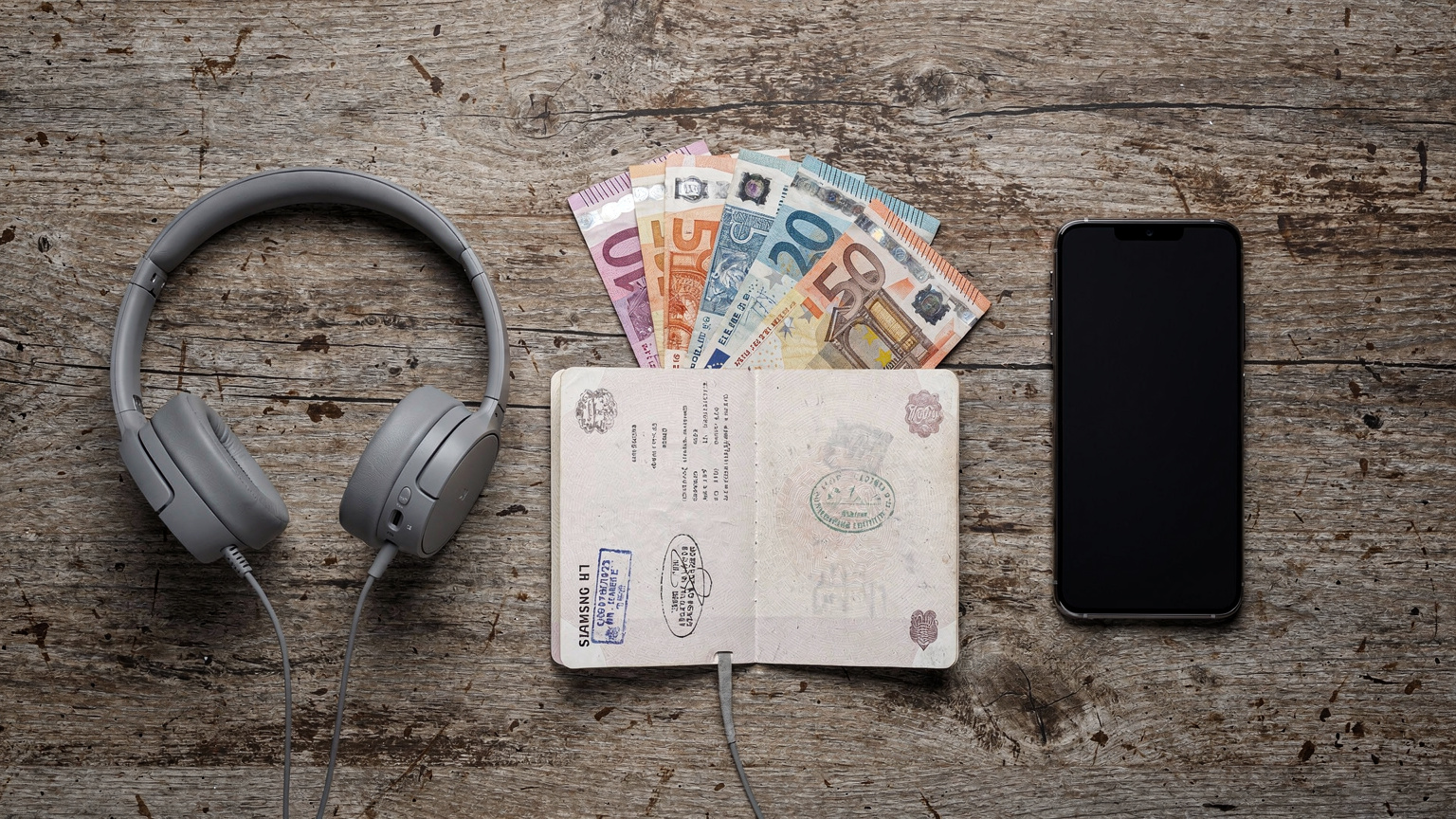 O ilustrație detaliată a diferitelor monede, inclusiv lei românești și euro, așezate ordonat lângă un pașaport deschis, un Samsung Galaxy S26 și o pereche de căști minimaliste, pe o masă din lemn, în stil flat-lay, cu o lumină moale, de studio.
