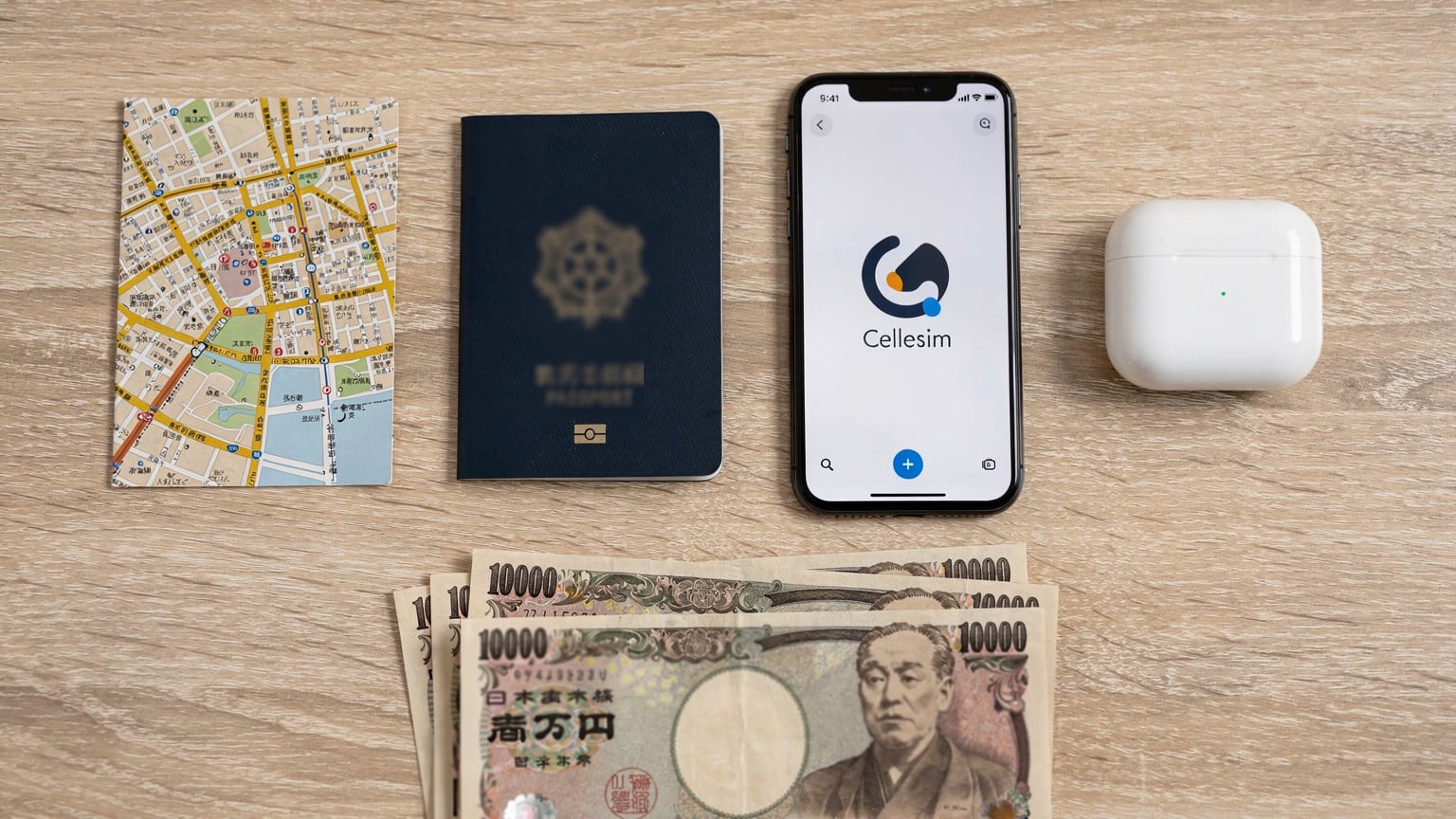 An overhead flat-lay of travel essentials: a passport, a smartphone showing a Cellesim logo, wireless earbuds, a small map, and Japanese Yen on a light wooden table.