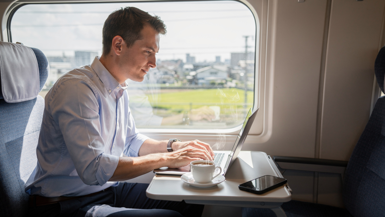 一名身著商務休閒裝的男士坐在東京新幹線列車的窗邊, 專心致志地使用筆記型電腦工作, 旁邊放著一杯咖啡和他的手機, 窗外是飛馳而過的城市風景。