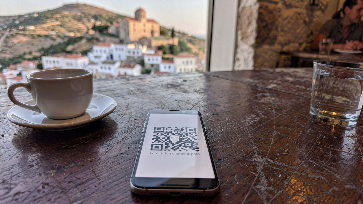 קוד QR של eSIM המוצג על מסך סמארטפון, ממוקם על שולחן עץ בבית קפה יווני עם רקע מטושטש של מנזר יווני עתיק וכפר ציורי.