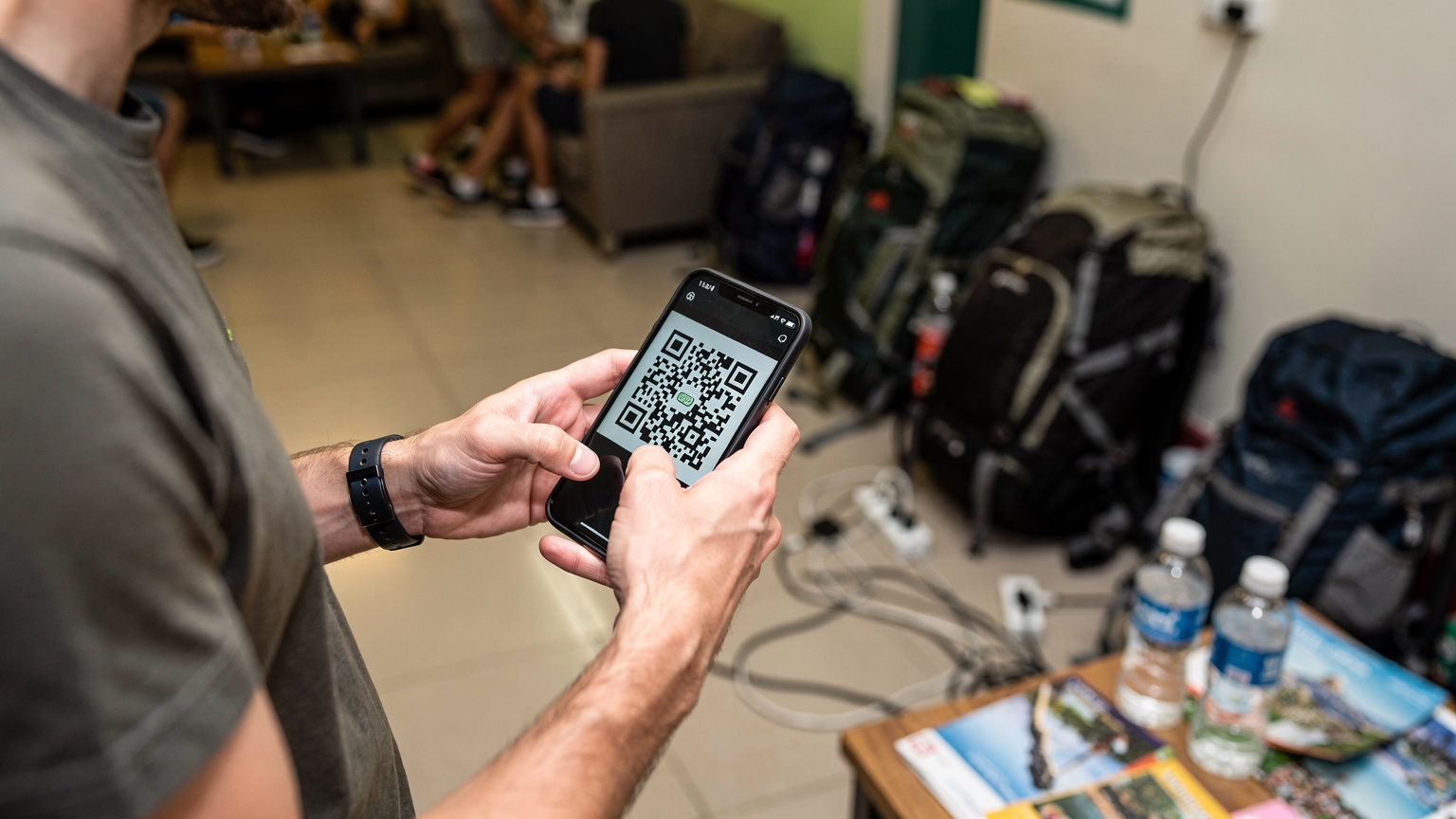 Un călător folosește o aplicație de eSIM pentru a scana un cod QR și a-și activa planul de date, pe un telefon modern, într-un hostel aglomerat din districtul Sukhumvit, Bangkok, cu alte rucsacuri și hărți în fundal.
