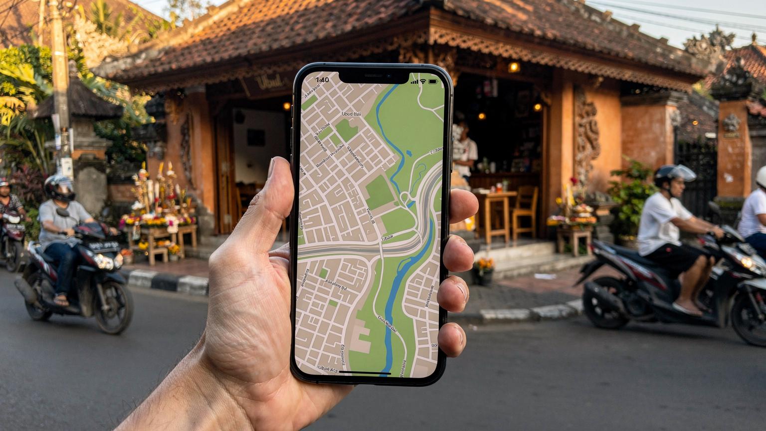 O mână umană ține un smartphone care afișează o hartă detaliată a cartierului Ubud din Bali, cu o cafenea locală și o stradă animată cu scutere indoneziene în fundal, la lumina dimineții.