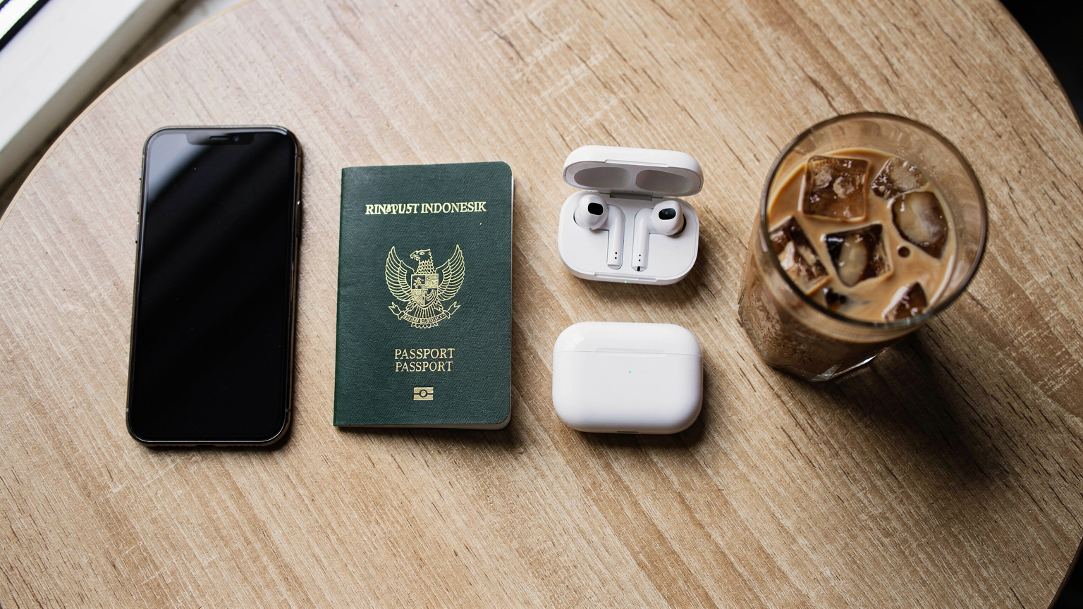 Sebuah flat-lay meja kafe dengan paspor Indonesia, ponsel, headphone, dan segelas kopi.