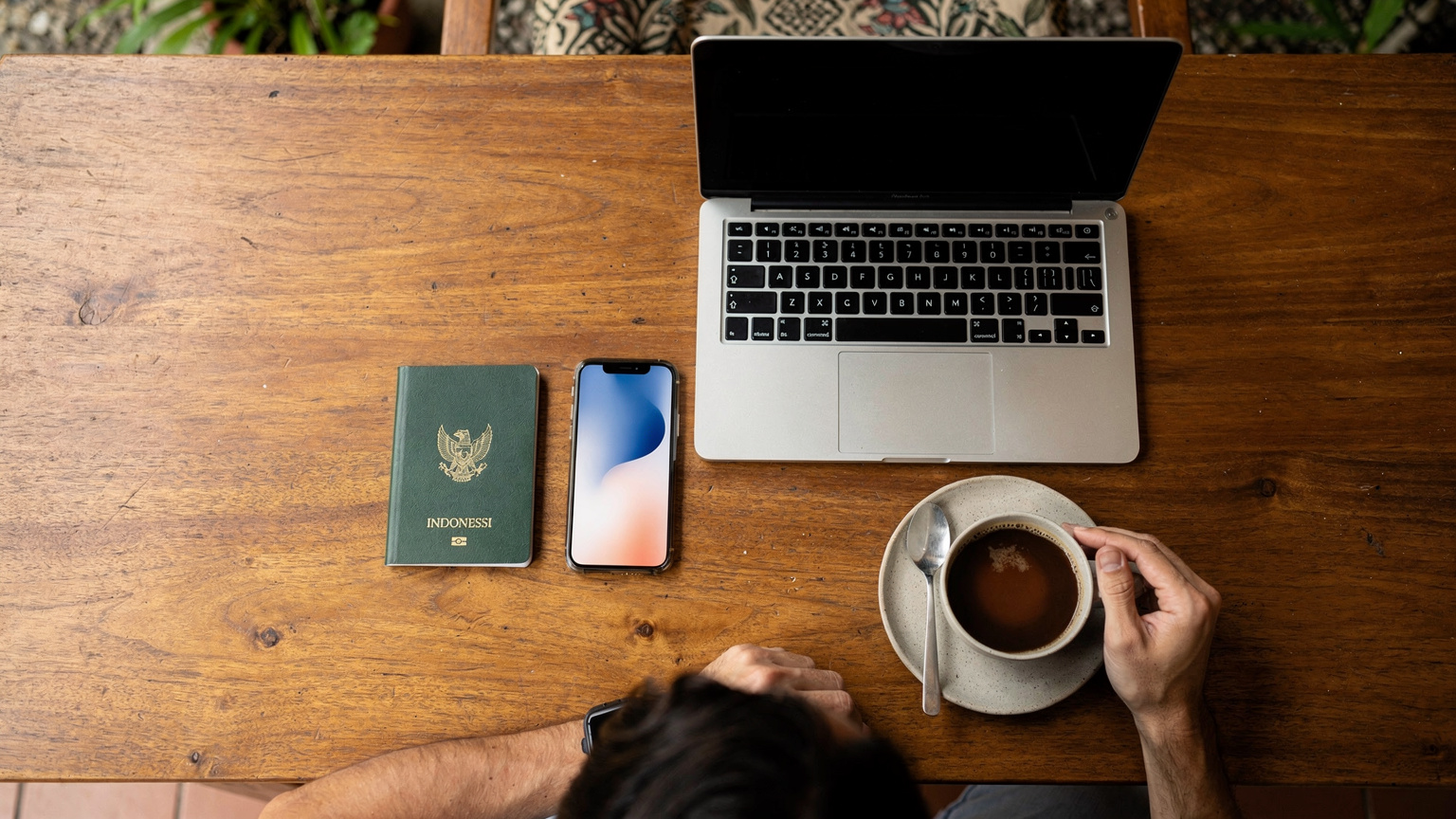 Tampak atas meja kerja yang rapi dengan paspor Indonesia, ponsel pintar yang menampilkan layar pengaturan eSIM, sebuah laptop, dan secangkir kopi, dengan nuansa tropis di latar belakang.