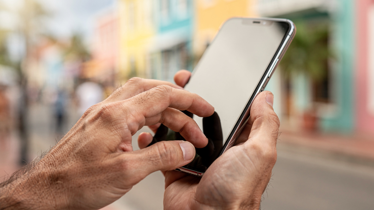 Close-up van handen die een smartphone vasthouden. De persoon tikt op het scherm, mogelijk een kaart-app of social media, met op de achtergrond een vaag zicht op een kleurrijke Curaçaose straat.
