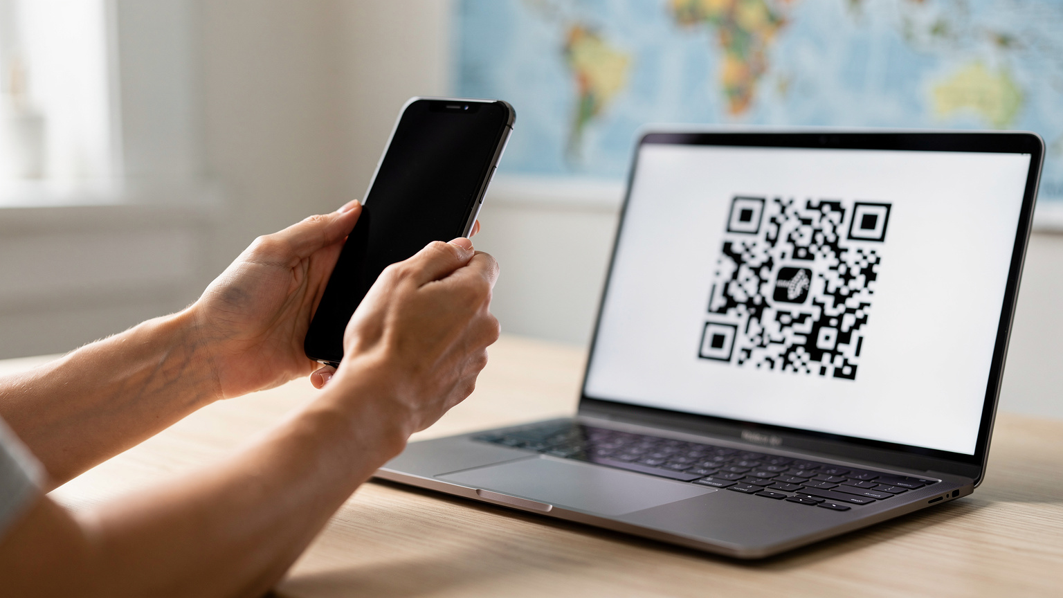 Zblízka, ženské ruky držia iPhone a skenujú QR kód eSIM z obrazovky notebooku, v pozadí je rozmazaná mapa sveta.